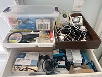 Overview of storage cabinet with magnifying glass, plastic box with paper clips, tape, and wooden tray holding cables and devices.