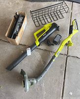 Overview of Ryobi string trimmer, leaf blower, battery, and charger arranged on garage floor with a box and grill rack nearby.