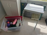 Overview photo showing clear plastic bin with colorful yarn skeins, white portable adjustable table, and quilting mats stacked behind the table.