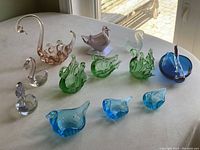 View of 12 assorted glass swans in clear, amber, purple, green, and blue on white surface near window with sunlight.