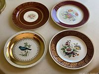 Four decorative plates featuring two with floral center designs and two with bird motifs and gold accents in ornate borders.