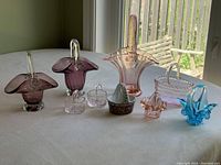 Photo showing all 9 decorative baskets arranged on a table near a window for natural light. Includes various glass baskets in pink, blue, amber, clear, and one ceramic basket.