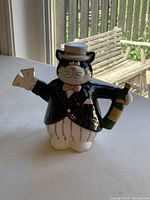 Front view of the cat shaped ceramic teapot showing it holding a champagne bottle and a horn, also wearing a tuxedo jacket, pink bow tie and white hat.
