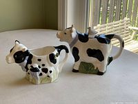 Photo showing ceramic black and white spotted cow teapot and smaller matching creamer on table by window