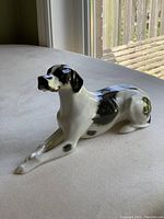 Side angled photo of the porcelain English Pointer dog figurine on a grey surface near a window, clearly showing black and white coloration and glossy finish.