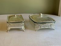 Two rectangular glass serving dishes in silver-tone metal holders with lids, showing the intricate design of the holders and matching lids, viewed from front and side angles.