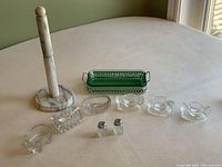 Photo of the full lot on a table showing a marble paper towel holder, green glass pickle/butter dish with metal handles, three clear glass hen egg cups, three assorted salt cellars including one with a scoop, and two mini salt and pepper shakers.