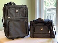 Matching set of Outline brand carry-on luggage and hand tote bag placed on a surface near a window, showing their size and appearance together.