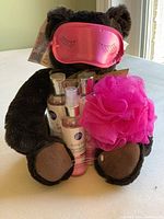 Brown stuffed teddy bear holding three bottles of Miss Spa branded bath & body products and a bright pink netted shower sponge, all arranged on a surface near a window.