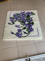 Top-down view of the square shaggy carpet showing purple flower and green leaf pattern on cream background with purple corner borders. Carpet texture is thick and plush.