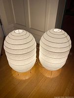 Two larger white Ikea Storuman paper lantern lamps illuminated on wooden floor, shown side by side.