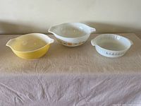 Three vintage mixing bowls set side by side on a cloth surface: yellow Pyrex, white Pyrex with Town and Country pattern, and small white Fire King bowl with Candle Glow pattern.