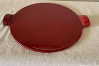 Red circular pizza stone with smooth glazed surface and two handles on sides, placed on fabric surface.
