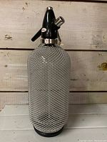 Full view of Kovocas soda siphon showing metal mesh covered glass bottle with black plastic head and chrome spout.