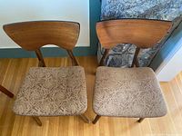 Front view of two mid-century wooden dining chairs showing curved backs and beige patterned upholstery on seats.