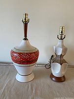Pair of vintage mid-century ceramic lamps on a table background. Larger lamp has floral and geometric reddish-orange patterns; smaller lamp has abstract geometric design with ribs and wooden base.