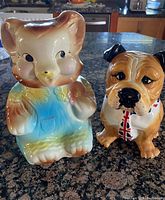 Front view of the two ceramic cookie jars on a granite counter: colorful bear with bib on the left and American Bulldog with bib on the right.