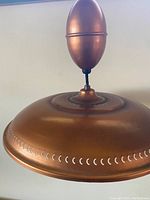 Close-up side view of large copper light fixture showing curved dome shade with perforated crescent-shaped pattern around edge and rounded finial on top.