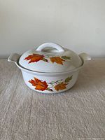 Front view of the vintage enamel casserole with lid displaying autumn leaf decoration, cream background, and side handles.