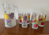 A full view of the glass jug with handle and spout alongside six matching tumblers, all decorated with colorful painted fruits.