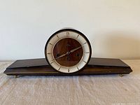 Full frontal view of the vintage German mantel clock showing wood and metal construction with a long base and round clock face, missing glass front.