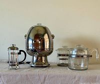 Full view of all four coffee makers: metal coffee urn, French press, two glass stove top coffee makers, all placed on a beige cloth.