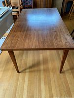 Full view of the teak veneer dining table showing rectangular top and tapered legs against wood floor background.