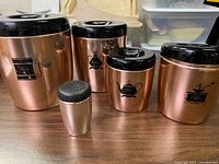 Front view showing all 5 items with rose colors and black lids, canisters labeled Flour, Coffee, Sugar, Tea, and a shaker.