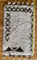 Top view of the small rectangular wool rug with a measuring tape indicating length, showing abstract brown diamond and triangle pattern on cream background and knotted edge fringe tassels.