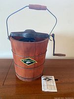 Front view of wooden bucket ice cream maker with metal crank lid and instruction booklet nearby