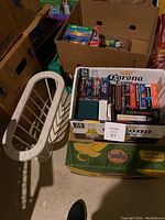 Boxes of assorted books, audio books on CDs and cassettes set beside a white metal magazine rack