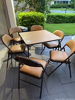 Full view of the square card table surrounded by six matching cushioned chairs placed outdoors on a paved surface with greenery in the background.