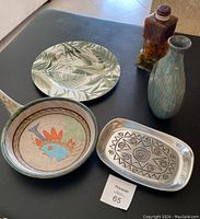 Five items arranged on a black surface: Mikasa tropical leaf platter, decorative Portuguese pottery fry pan with a colorful fish motif, Wilton silver-tone rectangular plate with black stars and spirals, green single flower vase, and a decorative decanter filled with peppers in oil.