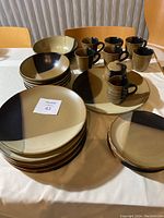 Photo showing stacks of dinner plates, dessert plates, mugs, serving bowl, serving platter, cream pitcher, and covered sugar bowl all in matching tri-color black, brown, and cream ceramic