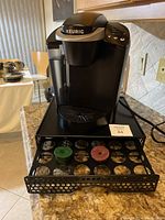 Black Keurig coffee machine on kitchen countertop with black metal pod holder drawer extended, two refillable pods visible.