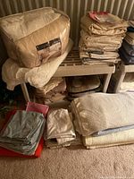 Photo showing a stack of packaged and folded bed linens including sheet sets and blankets in beige and neutral colors on a plastic shelf.