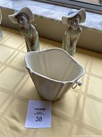 Two Lladro twin girl figurines posed with hands on knees along with a cream-colored short wide ceramic vase with scalloped rim and handles on a yellow tablecloth.