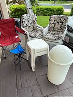 Four padded plastic chairs with floral cushions, red portable camping chair, white plastic small tables and white plastic trash can with swing lid on outside patio area.