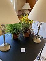 Two silver table lamps with white shades and two artificial plants on a table