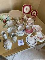 Full set of assorted tea cups, saucers, cream and sugar sets, decorative plates, Lenox cake platter, and bud vase arranged on a mirrored surface.