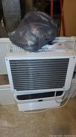 Photo of General Electric air conditioner unit with a grey plastic bag and papers on top, showing the front grille and controls.