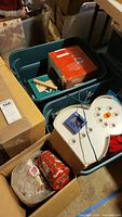 Photo showing two green plastic bins filled with various boxed Christmas decorations, including decorative tins and a snow globe.