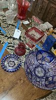 Overview of cut-to-clear colored crystal and art glass items on wooden table including cobalt blue bowl, cranberry pitcher, vase, and square dish
