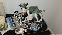 Vintage Fitz & Floyd Heidi Holstein black and white ceramic cow tureen with three accompanying cow salt, pepper, cream, and sugar containers, artisan duck decoys, and corncob pipe arranged on black glass table.