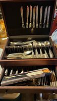 Full view of open wooden storage box with knives in upper lid rack and forks, spoons, and other flatware in lower compartments with an additional drawer containing more cutlery and boxed item.