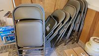 Stack of 10 Cosco vinyl padded metal folding chairs leaning against wall in corner on tile floor. Chairs have beige vinyl padded seats and backs with matching metal frames.
