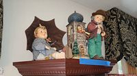 Two Hummel figurines on display shelf: a boy figure next to a small castle and a girl figure with umbrella sitting down.