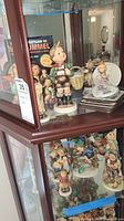 Full view of glass display cabinet containing multiple Hummel Goebel figurines and a collector's guide book.