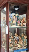 Wide view of multiple Hummel Goebel figurines arranged on a glass shelf inside a wooden cabinet, showcasing detailed hand-painted ceramic figures resembling children in various poses.