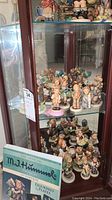 Photo showing multiple Hummel porcelain figurines arranged on glass shelves inside a cabinet plus visible pricing tag on door